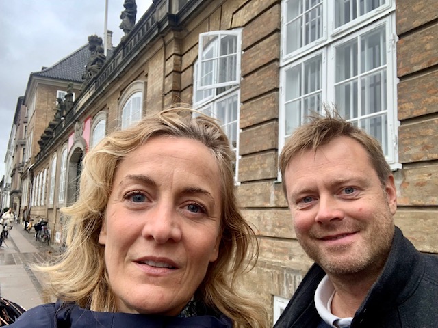 Et billede af to personer, en kvinde( Anna Foss) og en mand (Rasmus Aude), der tager en selfie. Kvinden er forrest i billedet med blondt, bølget hår og en mørk jakke, og hun smiler bredt. Manden bag hende har kort, lyst hår, er iført en mørk jakke over en lyseblå skjorte og smiler. De står udendørs foran Nationalmuseet, og der ses cykler og en åben dør i baggrunden.