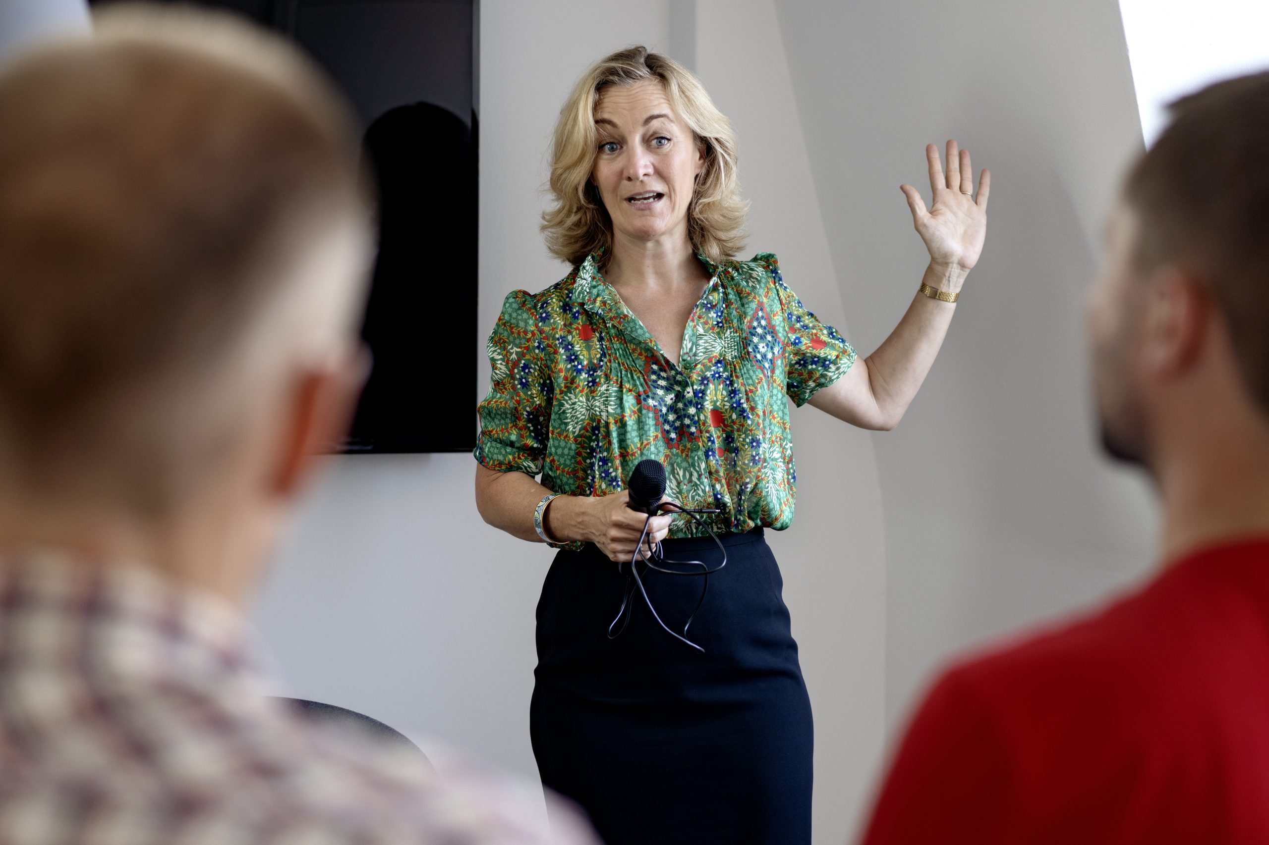 Billedet viser medietræner Anna Foss midt i en session om budskabsudvikling. Hun er klædt i en farverig bluse, står smilende og holder en mikrofon, klar til at guide en person i forgrunden, som kun er delvist synlig. Baggrunden er en professionel indstilling med studielys, hvilket tyder på en praktisk øvelse i medietræning, hvor fokus er på at forbedre pressemeddelelsekommunikation og pressehåndtering. Dette indikerer et levende scenarie, hvor kommunikationsstrategier og budskabstræning er i centrum.