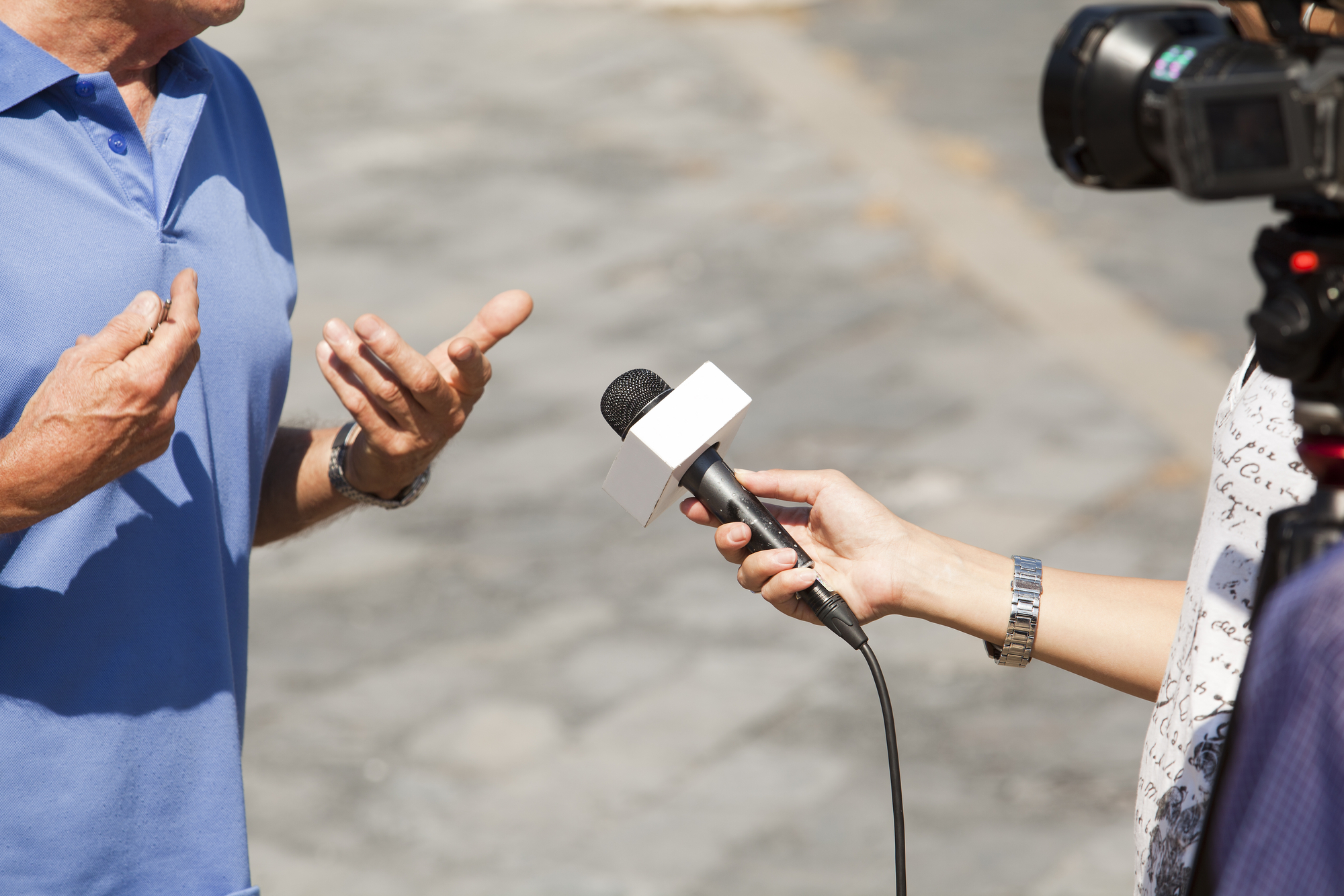 En reporter med et mikrofonholder med en blank plads, hvor et logo kan indsættes, interviewer en person i en blå poloshirt. Personen gestikulerer med sine hænder og ser ud til at forklare noget. Baggrunden er sløret, men det ser ud til at være et udendørs miljø med brolagte sten. En videokamerafører optager interviewet.